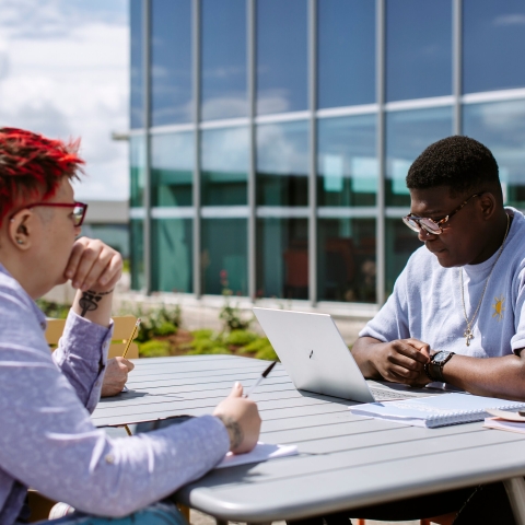 Picture of two people sat on a bench outside of a university building. They are bothlooking at laptop screens and working hard. 