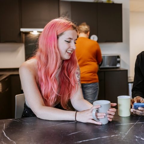 A female and male student are leaning on a kitchen counter with mugs smiling