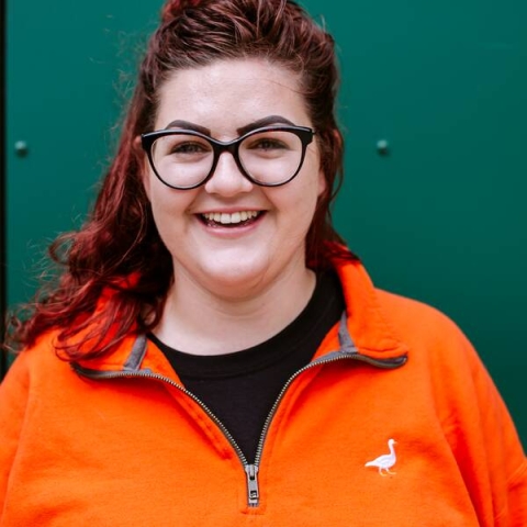 Student with brown hair and orange jumper standing in front of a green wall