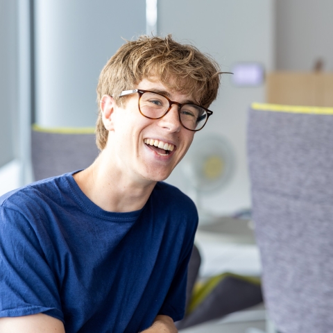 Male student in blue teshirt leaing on the arm on a chair while smiling