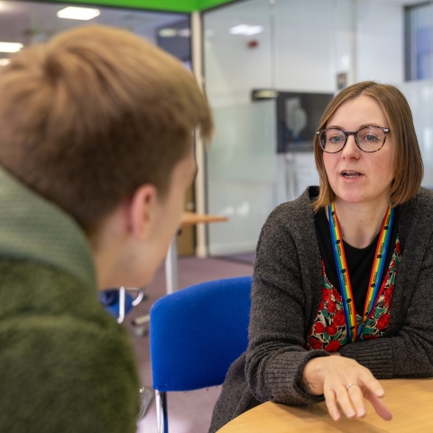 A staff member talking with a student at a table in a university learning space.