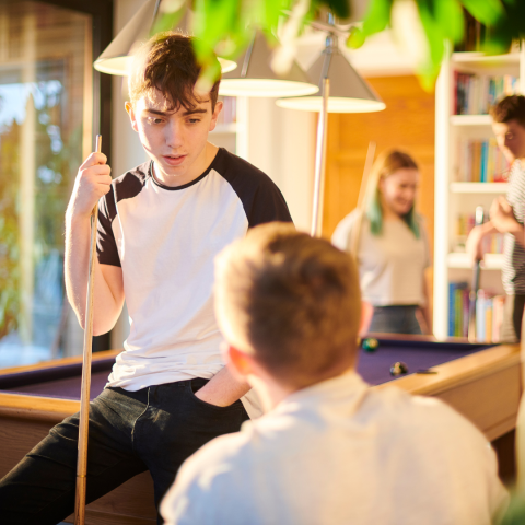 Young man holding a pool cue, concentrating on the game, with two people in the background at a home pool table.