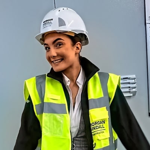 A woman in viz jacket and hard hat, standing in front of a whiteboard with writing on it, smiling at the camera