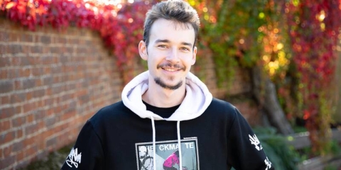 male student wearing a hooded sweatshirt standing in front of autumn trees