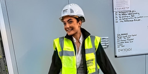 A woman in viz jacket and hard hat, standing in front of a whiteboard with writing on it, smiling at the camera