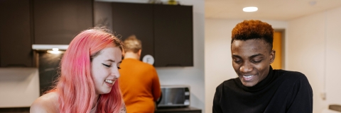 A female and male student are leaning on a kitchen counter with mugs smiling