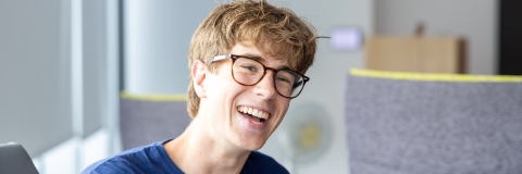 Male student in blue teshirt leaing on the arm on a chair while smiling