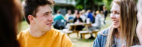 Students chatting at a picnic bench in the park, one wearing a yellow jumper and the other a denim jacket