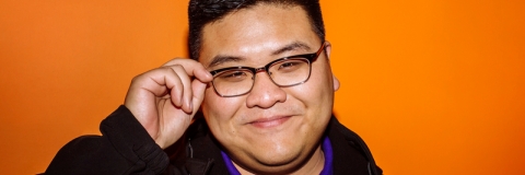 Smiling male student hold glasses in front of orange wall