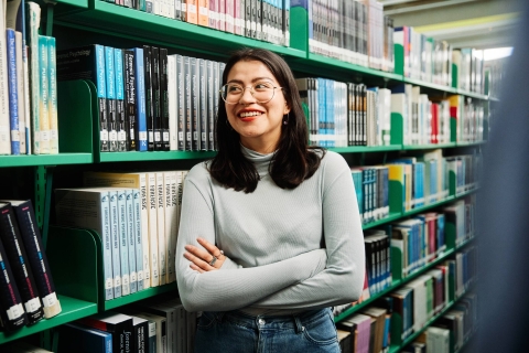 student with arms folded in front of bookshelf