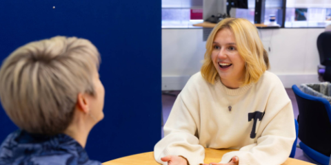 A student sat at a table with an adviser receiveing advice. They are deep in discussion and the student is smiling widely. 