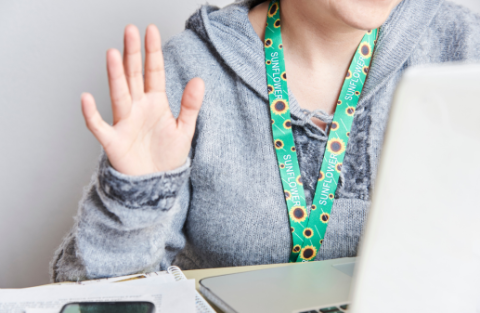 Woman in online meeting waving. She is wearing a sunflower lanyard which indicates she has a disability.