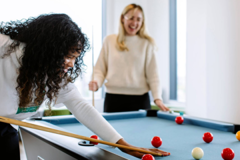 Two female students are playing pool, one in the foreground is set to strike the ball with a cue, the other is smiling in the background