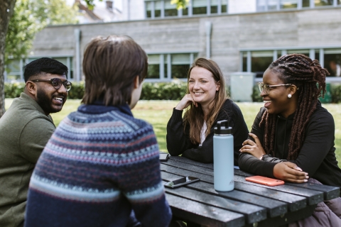 Four students of different ethnicities are sitting around a table talking and smiling.