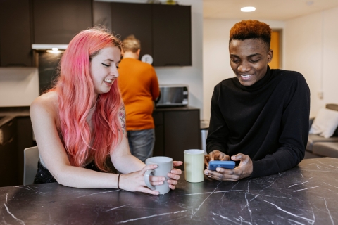 A female and male student are leaning on a kitchen counter with mugs smiling