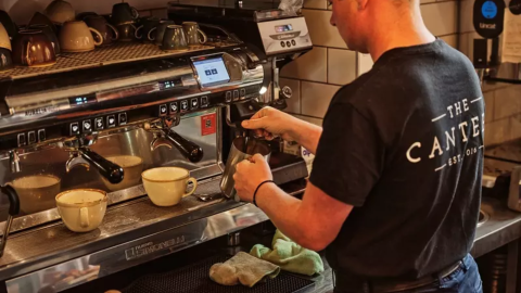 A person in a uniform working at a cafe. They are making coffee using a coffee machine.
