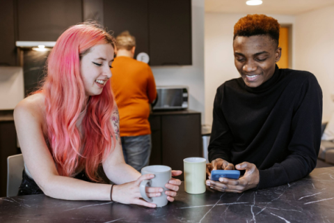 A male and female student are sat at a kitchen counter together with cups, the male student has a phone in hand and both are smiling.