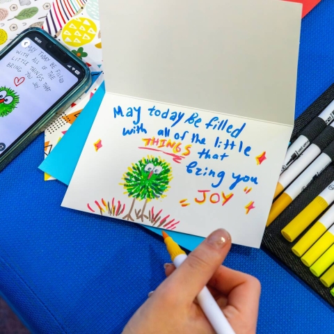 A mobile phone, colourful pens and a drawing in a card upon a table. The card says 'May today be filled with all of the little things that bring you joy'