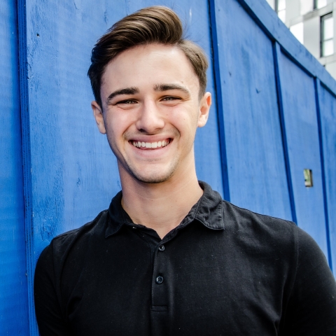 Josh Robinson smiling to camera wearing black t-shirt against blue wall