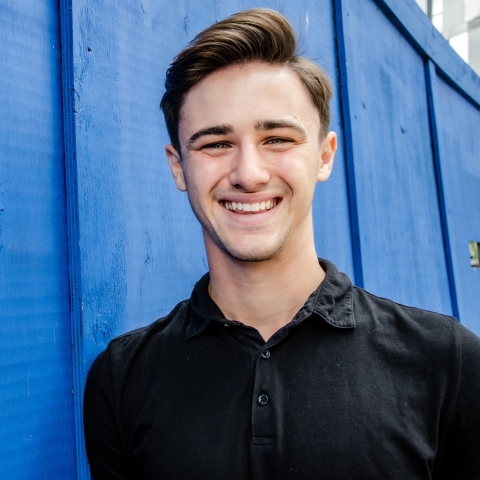 Josh Robinson smiling to camera wearing black t-shirt against blue wall