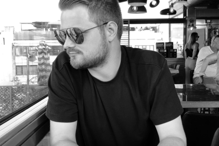 Black and white photo of Kieran Graham, sat in a restaurant wearing sunglasses and looking out the window.