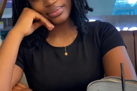 Female student sitting at a table. Wearing black tshirt and shoulder length hair. Behind her is a lamp and in front on the table is a bag, cup and glasses.